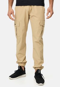 Pantalones cargo beige con puños elásticos, dos bolsillos laterales con solapas, fabricados en un tejido ligero y combinados con zapatillas deportivas negras.