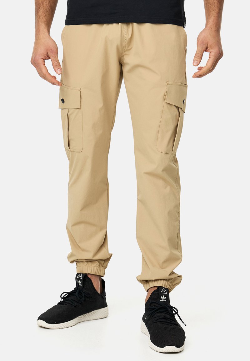Pantalones cargo beige con puños elásticos, dos bolsillos laterales con solapas, fabricados en un tejido ligero y combinados con zapatillas deportivas negras.