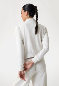Felpa bianca a cropped con tessuto testurizzato, polsini a costine e strisce laterali. Indossata con pantaloni testurizzati abbinati, mostrata da dietro.