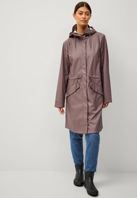 Chubasquero impermeable color malva con capucha, cintura con cordón y dos bolsillos frontales, combinado con jeans azules y botas de goma negras.