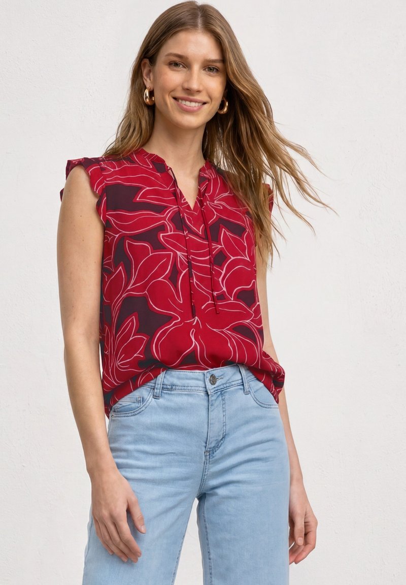 Jeune femme souriante portant un haut sans manches rouge à fleurs et un jean bleu clair sur un fond blanc uni.