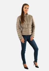 Veste matelassée légère couleur taupe avec des coutures verticales, col montant, et fermeture éclair, associée à un jean skinny noir et des escarpins noirs.