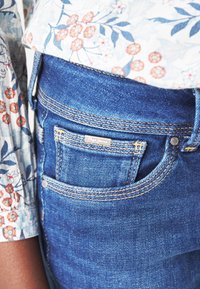 Jeans z modrého denimu s přiléhavým střihem mají charakteristick�é švy a malou značkovou nášivku na zadní kapse, doplněné květinovým vzorem košile.