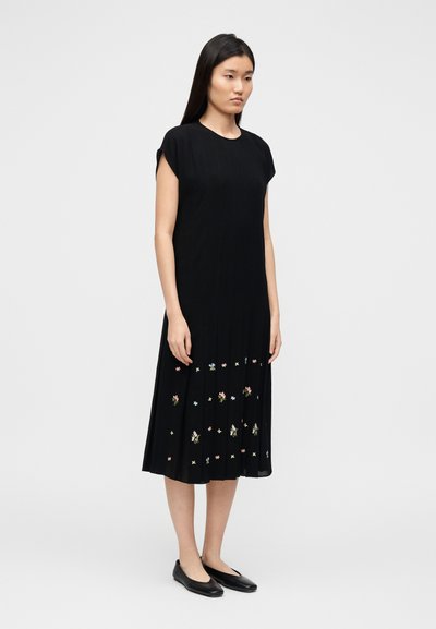Robe noire plissée à manches courtes, ornée de broderies florales sur l'ourlet. Tissu lisse avec une texture subtile et une silhouette fluide.