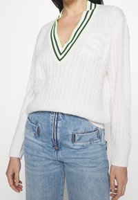Weißer Strickpullover mit tiefem V-Ausschnitt, ausgestattet mit einem schwarz und neon-grün gestreiften Kragen und einem gestickten Logo auf der Brust.