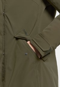 Oliven grøn vandtæt jakke med blød tekstur, der har knaplukning og sidelommer, fremhævet af en subtil logo-detail på ærmet.