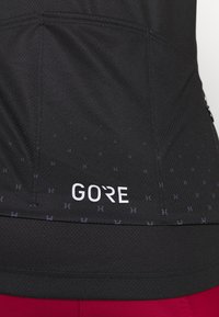 Camiseta deportiva negra con tejido texturizado, un patrón sutil y un prominente logo blanco de "GORE" cerca del dobladillo.