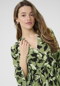 Grünes Kleid mit V-Ausschnitt und langen Ärmeln, das ein mutiges abstraktes Muster in Schwarz und Hellgrün aufweist und einen schwingenden Saum hat.