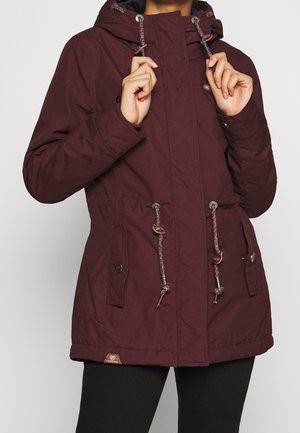 Femme ajustant les cordons de la capuche d'une veste à capuche violet foncé avec des poches à boutons et des cordons à l'avant.