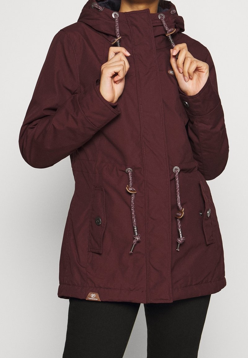 Femme ajustant les cordons de la capuche d'une veste à capuche violet foncé avec des poches à boutons et des cordons à l'avant.