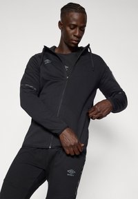 Čierna bunda na zips s kapucňou, s hladkým povrchom, trendy strihom a decentným označením značky. V kombinácii s čiernymi nohavicami a detailnými akcentmi.
