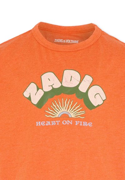 T-shirt orange à manches courtes avec le texte audacieux et stylisé « ZADIG », un graphique de soleil au-dessus, et « HEART ON FIRE » en texte bleu en dessous.