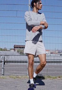 Maglietta sportiva a maniche lunghe grigia chiara, pantaloni corti bianchi e scarpe da ginnastica nere. L'individuo è in piedi accanto a una recinzione metallica in una giornata di sole.