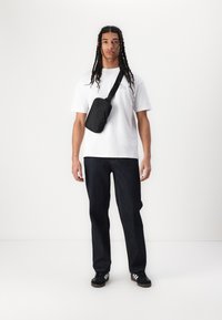 Svart korsbåndveske med glidelås brukes over en hvit T-skjorte og mørk denim bukser. Modellen står vendt mot kamera med et nøytralt uttrykk.