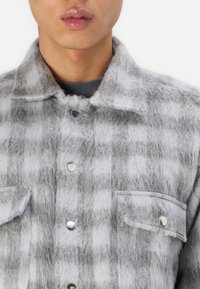 Grijs en wit geruit flanellen shirt met een fuzzy textuur, voorzien van twee borstzakken en zilveren knoopsluitingen.