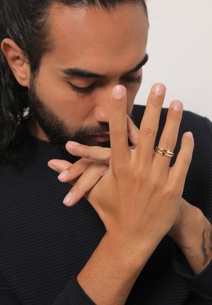 Gold ring with intersecting lines on the right hand. Skin tone is light brown. Hand poses thoughtfully with fingers clasped. Dark navy shirt texture visible.