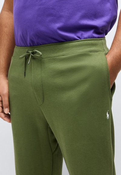 Άτομο που φοράει χακί παντελόνι με κορδόνι και λευκό λογότυπο πόλο, με πουκάμισο μωβ μπουκαρισμένο μέσα, τα χέρια σταθερά στις τσέπες.