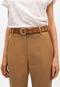 Ruskea nahkavyö, jossa on kultaiset osat ja niittiyksityiskohdat, käytetty vaaleiden housujen ja valkoisen paidan päällä, jossa on vaaleanruskea brodeeraus.