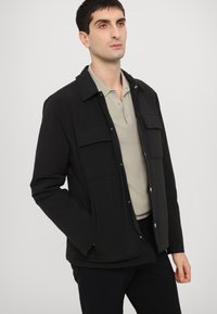 Jachetă exterioară neagră cu guler, două buzunare frontale și butoane cu capse, purtată peste un tricou polo gri deschis. Textură netedă.