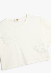 Camiseta de algodón en un tono blanco roto con un corte relajado, que presenta dos diseños de corazones de encaje bordados en la parte delantera, mangas cortas y cuello redondo.