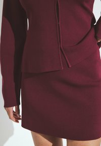 Burgundy-neulepaita pitkillä hihoilla, jossa on kerroksellinen muotoilu ja sidottu yksityiskohta. Tämä asu täydentää yhteensopiva kapea hame.