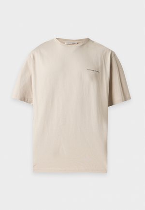 Homme debout, les mains dans les poches, portant une chemise beige à manches courtes, un pantalon cargo foncé et des baskets noires sur fond blanc.