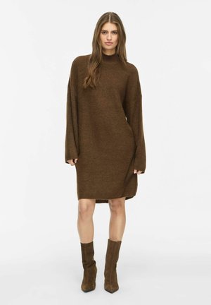 Woman standing wearing a loose brown long-sleeve sweater dress with matching brown suede boots against a white background.