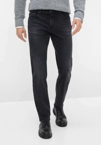 Jeans neri in denim con vestibilità slim, dotati di tasche frontali e chiusura a bottone, abbinati a scarpe in pelle nere e un maglione grigio in maglia.