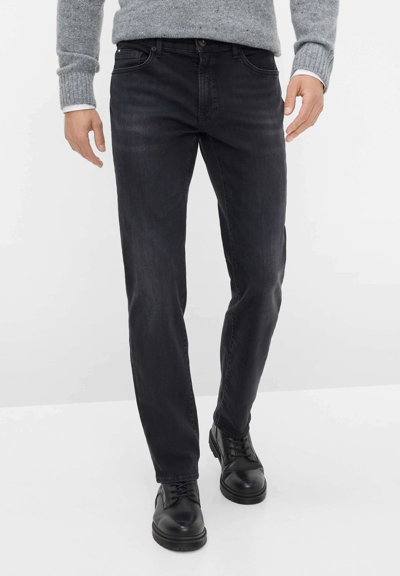 Jeans neri in denim con vestibilità slim, dotati di tasche frontali e chiusura a bottone, abbinati a scarpe in pelle nere e un maglione grigio in maglia.