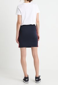 Gonna blu navy con strisce rosse sui lati, abbinata a una t-shirt bianca, indossata con sneakers nere. Tessuto morbido, forma aderente, design casual.