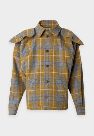 Homme portant une veste à carreaux moutarde et grise par-dessus une chemise blanche à boutons, pantalon beige et mocassins marron foncé, debout les mains dans le dos.