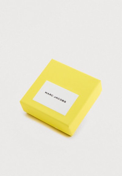 Gele cadeauverpakking met een glanzende afwerking, voorzien van een wit label met "MARC JACOBS" in zwarte tekst, gecentreerd aan de voorkant. Vierkante vorm.