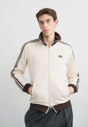 Fiatal férfi krémszínű sportdzsekit visel barna csíkokkal, barna gallérral és mandzsettákkal, kezeit a zsebében tartva, egy egyszerű világos háttér előtt áll.