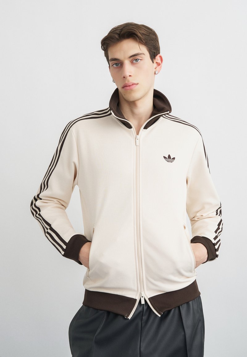 Joven con chaqueta deportiva crema con rayas marrones, cuello y puños marrones, manos en los bolsillos, de pie contra un fondo claro y liso.