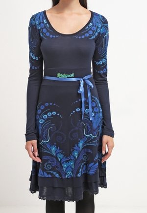 Femme portant une robe ajustée bleu marine à manches longues, ornée de motifs floraux bleus, avec une ceinture bleue nouée à la taille.
