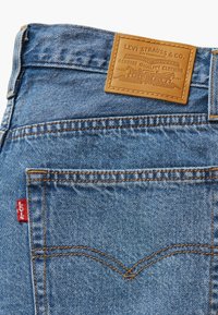Modre jeans iz denima z bež usnjenim označevalnikom z embosiranim blagovno znamko, oranžnim šivanjem in rdečim Levi's oznako na zadnjem žepu.