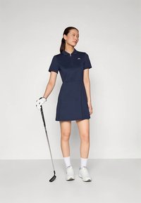 Φόρεμα γκολφ σε ναυτικό μπλε με λαιμόκοψη γιακά και κοντά μανίκια, με λεπτομέρεια φερμουάρ και λογότυπο στο στήθος. Συνοδεύεται από ένα μπαστούνι γκολφ.