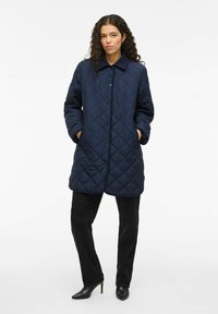 Mornarsko podloženo dolg plašč z ovratnikom, z gumbnim zapiranjem in stranskimi žepi. Nosi se preko črnih hlač in ostrih črnih čevljev.