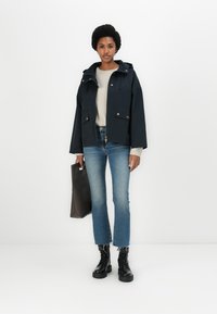 Veste courte bleu marine avec capuche, matériel doré, deux poches avant, pull crème, jeans bleus, bottes de combat noires et sac fourre-tout marron.