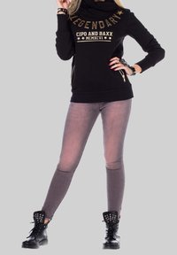 Sweatshirt noir avec inscription dorée, col drapé et poches zippées, associé à un jean skinny gris et des boots de combat noires cloutées.
