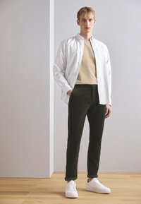 Chemise à rayures légères sur un t-shirt beige, associée à un pantalon vert foncé et des baskets blanches. Intérieur avec un sol en bois.