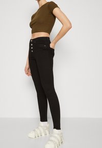Jeans skinny de cintura alta negros con detalles de botones plateados, combinados con una camiseta ajustada de manga corta en color verde oliva y zapatillas deportivas blancas.