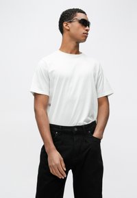 Biały t-shirt z krótkim rękawem w połączeniu z czarnymi spodniami, charakteryzujący się luźnym krojem i teksturowaną tkaniną. Model ma na sobie ciemne okulary przeciwsłoneczne.