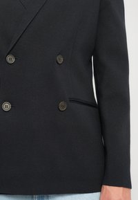 Tuvplāns, kurā redzams cilvēks, kurš valkā melnu divrindu žaketi ar četriem pogām un gaiši ziliem džinsiem.