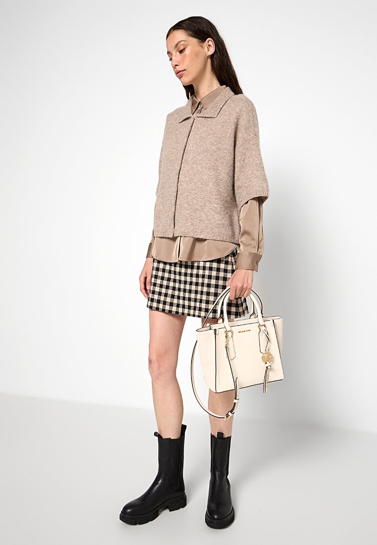 Beige Strickpullover, seidiges beiges Hemd, karierter schwarz-weißer Rock, schwarze kniehohe Stiefel und eine cremefarbene Handtasche mit Akzenten.