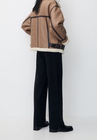 Giacca in faux shearling marrone con dettagli in pelle nera, foderata in bianco, e pantaloni larghi a righe nere a coste. Calzature nere eleganti completano il look.