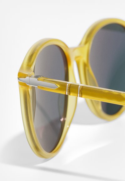 Lunettes de soleil jaunes avec une monture ronde, des verres teintés gris et un accent argenté sur les tempes. La monture a une texture translucide.
