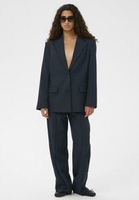 Sieviete ar gariem lokainiem matiem, ģērbta pārspīlēti lielā tumši zilā uzvalkā, melnās plakanās kurpēs un tumšos saulesbrillēs, stāv uz fona balta.