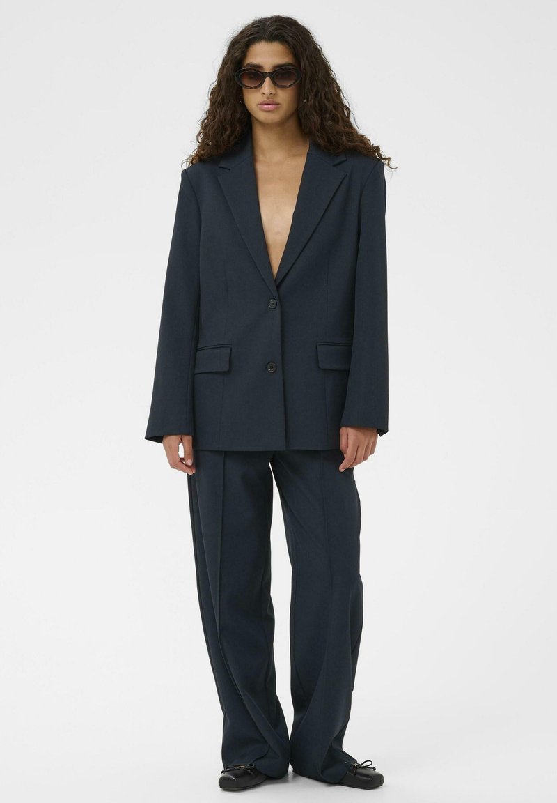 Sieviete ar gariem lokainiem matiem, ģērbta pārspīlēti lielā tumši zilā uzvalkā, melnās plakanās kurpēs un tumšos saulesbrillēs, stāv uz fona balta.