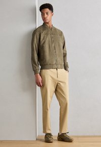 Olive green suede bomber jacket with ribbed cuffs, paired with beige chinos and olive sneakers. Model stands against a neutral background.